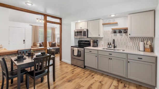 A modern kitchen with stainless steel appliances, a dining area with a wooden table and chairs, and a view of a living room with a sofa and curtains.