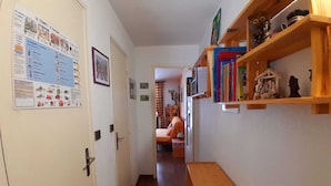 Un couloir étroit avec un banc en bois, une porte ornée d’un calendrier et une bibliothèque contenant des livres et des objets décoratifs.