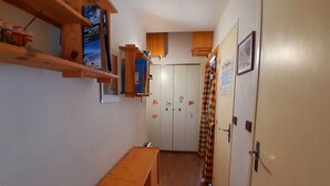 Un couloir étroit avec un banc en bois, une étagère remplie de livres et de bocaux, et une porte ornée d’un calendrier et d’autocollants.