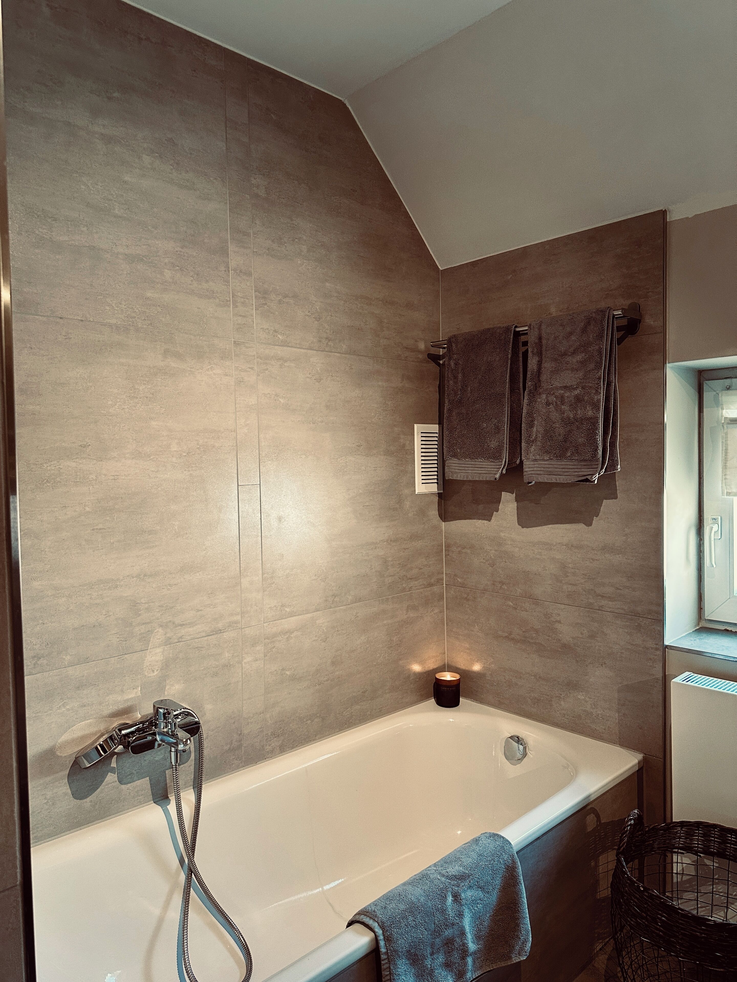 A modern bathroom with a bathtub, a towel rack with two towels, a small candle, and a wicker chair.