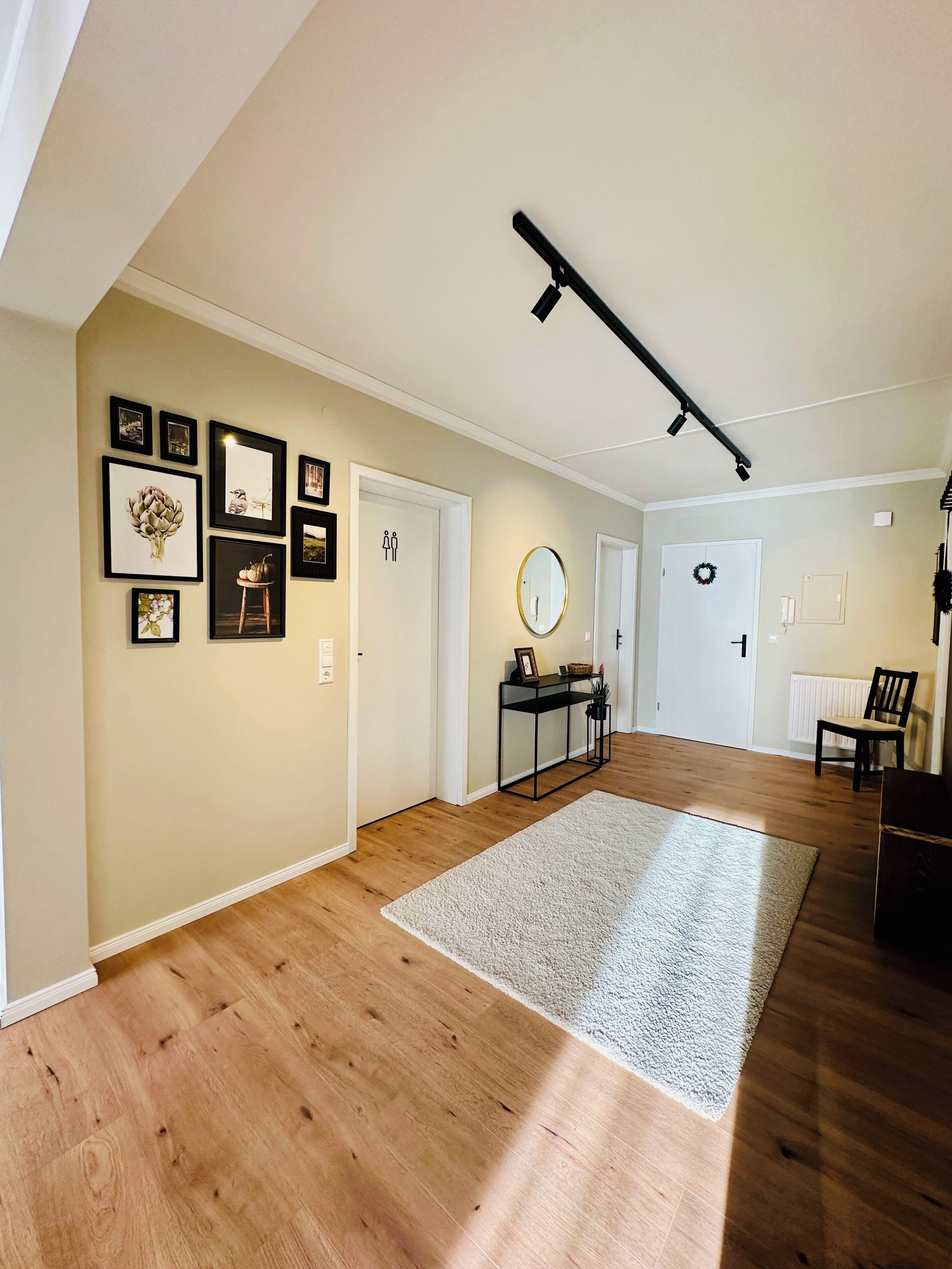 A spacious hallway with wooden flooring, framed artwork on the walls, and a central area with a round mirror and a small table.