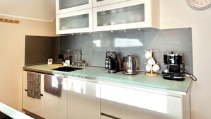 Une cuisine moderne avec des meubles blancs, un plan de travail en verre et divers appareils, notamment une machine à café et une bouilloire.