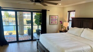 A hotel room with a large bed, a ceiling fan, a window with a view of a pool and buildings, and a nightstand with a lamp and a flower vase. - Be one of the first guests to enjoy this newly renovated luxury waterfront villa (Punta Gorda)