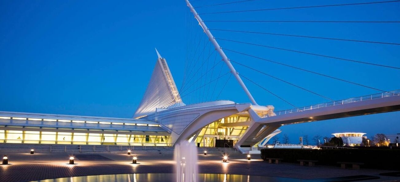 Un pont moderne au design original, caractérisé par une structure de câbles bien visible et un chemin illuminé menant à un bâtiment à la façade en verre.