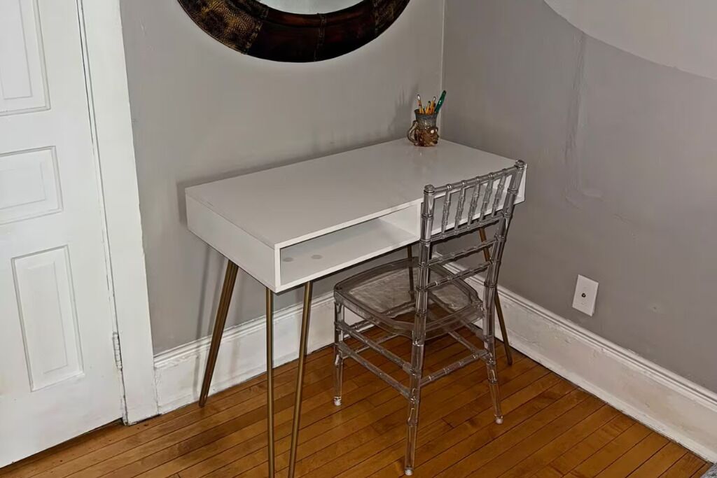 A white desk with a clear chair in a room with wooden flooring and a white door.