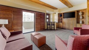 Amplia sala de estar con sofá, reposapiés y sillones, vigas de madera en el techo, televisor y puerta corrediza que da a un balcón.