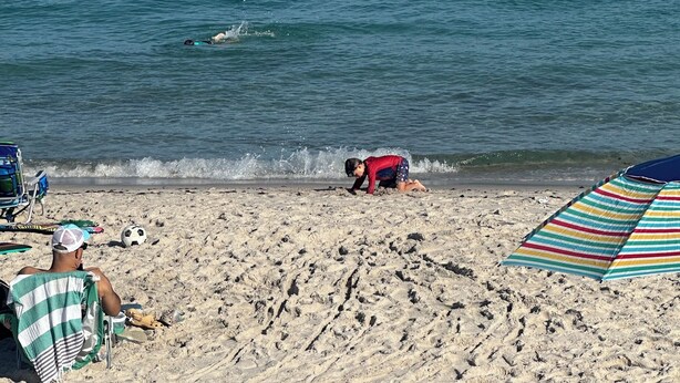 Una playa con arena, una sombrilla a rayas y una persona vestida de rojo.