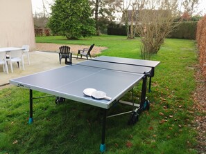 Een tafeltennistafel met twee batjes op een gazon, omringd door stoelen en bomen.