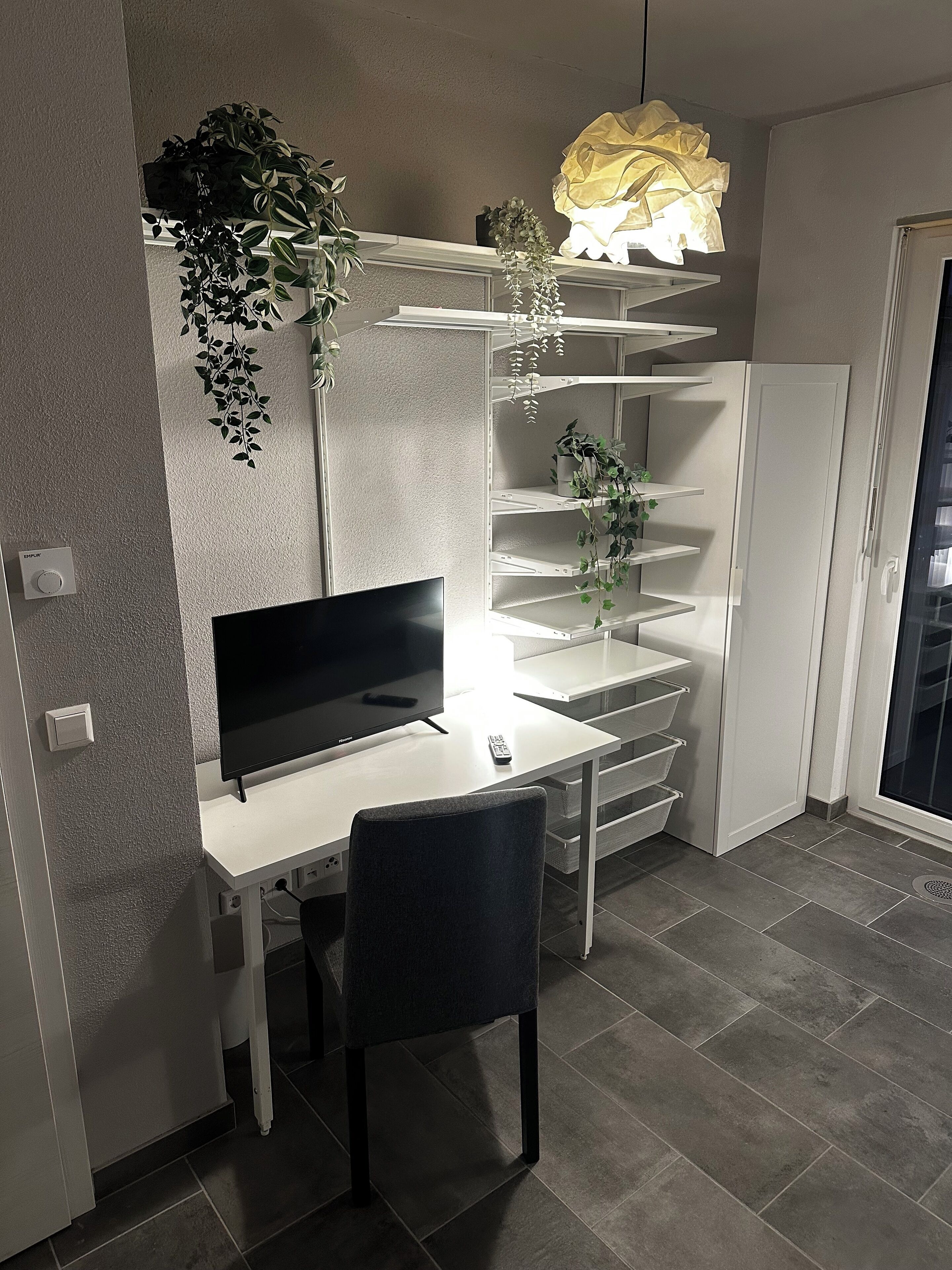 Un espacio habitable moderno y funcional, con un escritorio blanco, un televisor de pantalla plana, una silla y un estante con plantas.