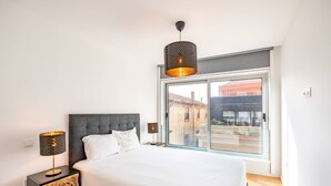 Ein modernes Schlafzimmer mit einem großen Bett, zwei Nachttischlampen, einem grauen Stoffpolster-Kopfteil und einem großen Fenster mit Blick auf Gebäude.