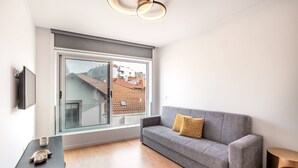 Ein modernes Wohnzimmer mit grauem Sofa, einem kleinen Tisch mit Dekoration, einem großen Fenster mit Blick auf Gebäude und einer einzigartigen, runden Leuchte.