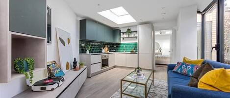 A modern kitchen with a central island, a dining area with a blue sofa, and a large window providing natural light.
