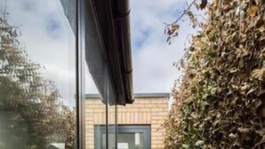 Un étroit sentier mène à une entrée en verre d’un bâtiment, lequel est entouré d’un mur de briques et d’une haie.