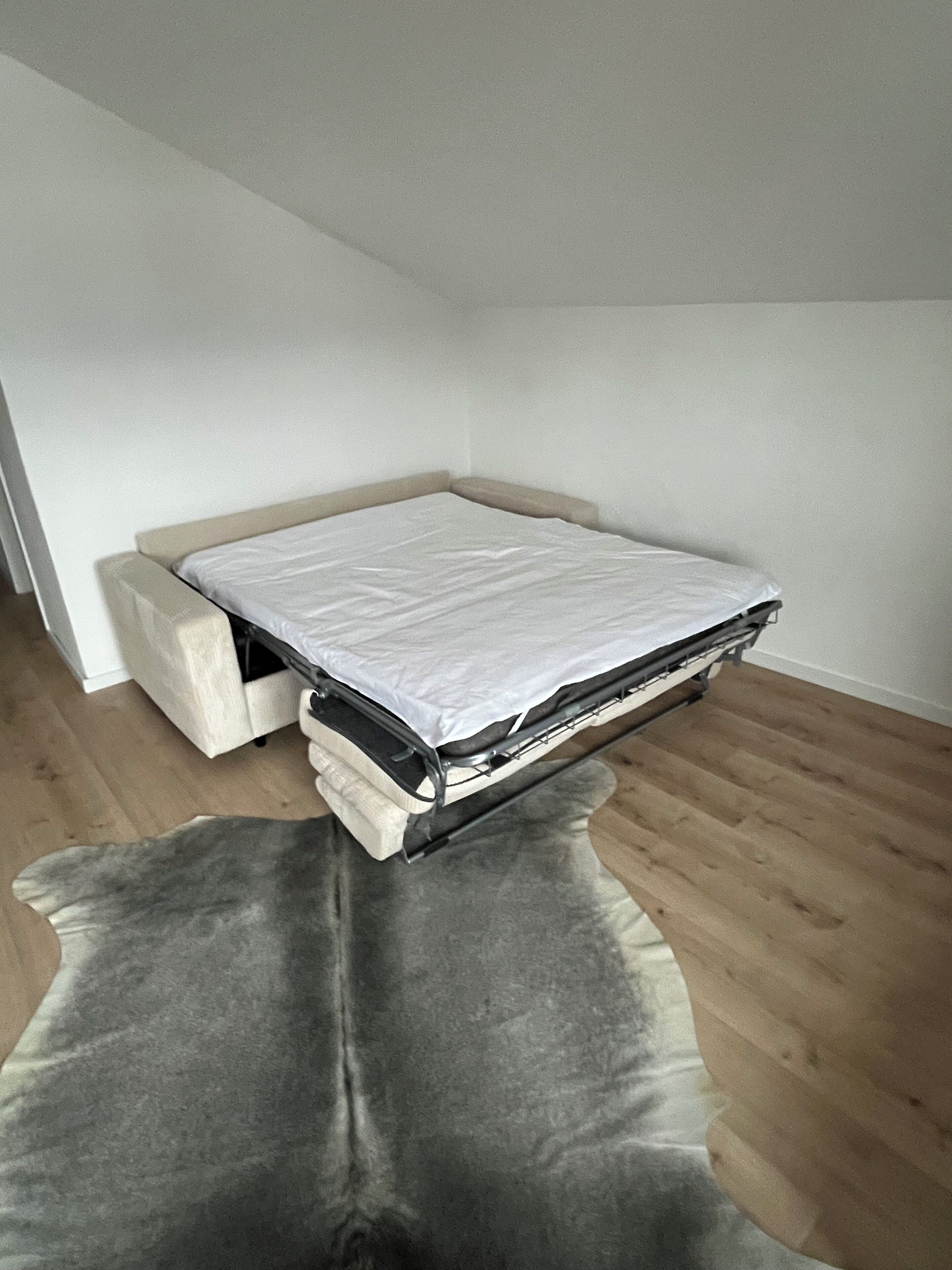 Ein kleines, kompaktes Bett in einem Eckzimmer mit Holzboden und einem grauen Teppich.