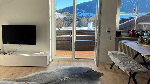 Ett modernt vardagsrum med en stor TV, en bekväm bänk, ett matbord och utsikt över ett snötäckt berglandskap genom en skjutdörr i glas.