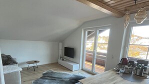 Ett modernt vardagsrum med ett träpanelat tak, ett stort fönster, en platt-TV, ett matbord och en matta i läder.