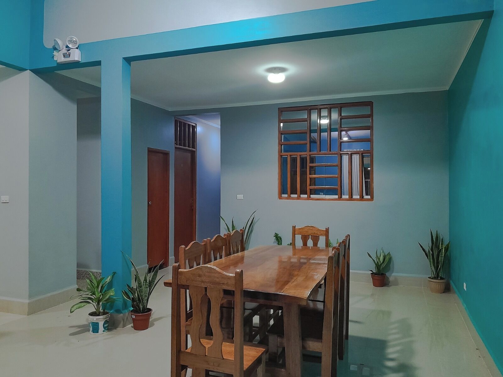 Un comedor con mesa y sillas de madera, plantas en macetas y una ventana con celosía de madera.