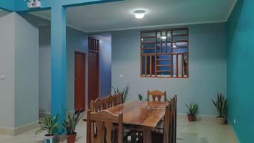 Un comedor con mesa y sillas de madera, plantas en macetas y una ventana con celosía de madera.