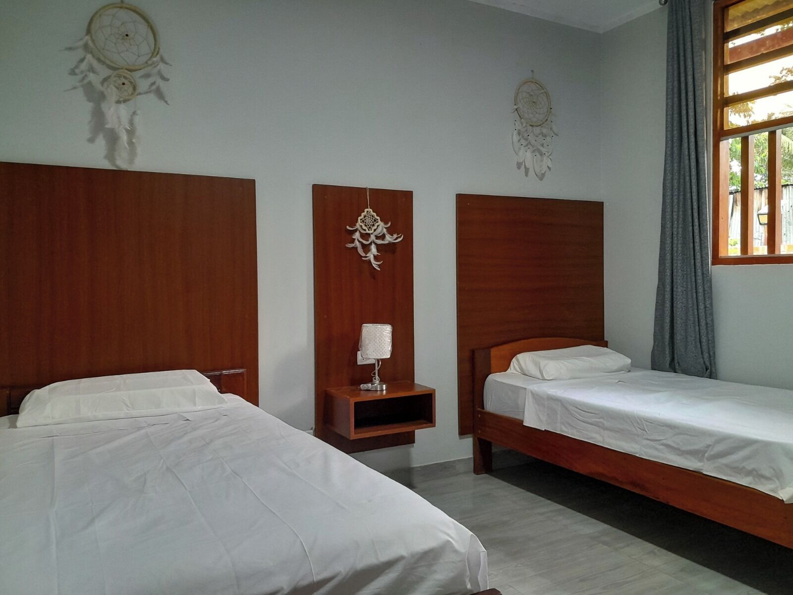 Ein Hotelzimmer mit zwei Einzelbetten, Holz-Kopfteilen, einem kleinen Nachttisch mit Lampe und einer Dekoration als Traumfänger.