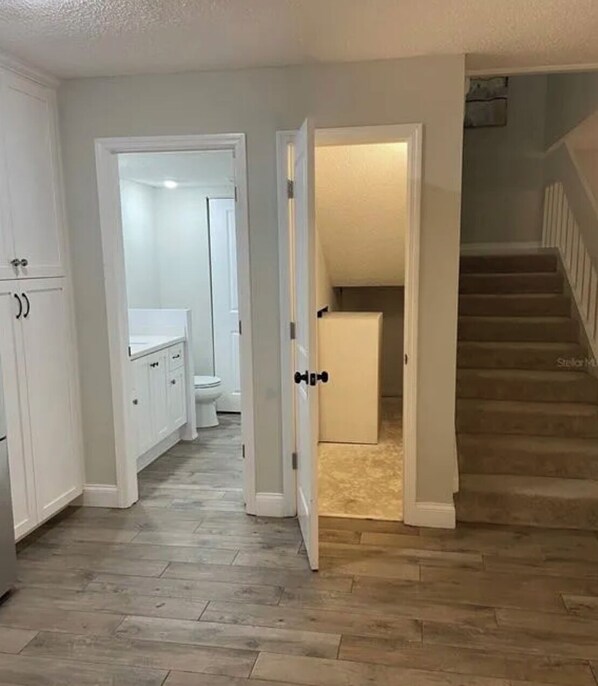A hallway with a wooden floor, white cabinets, a white toilet, and a staircase leading to a second floor.