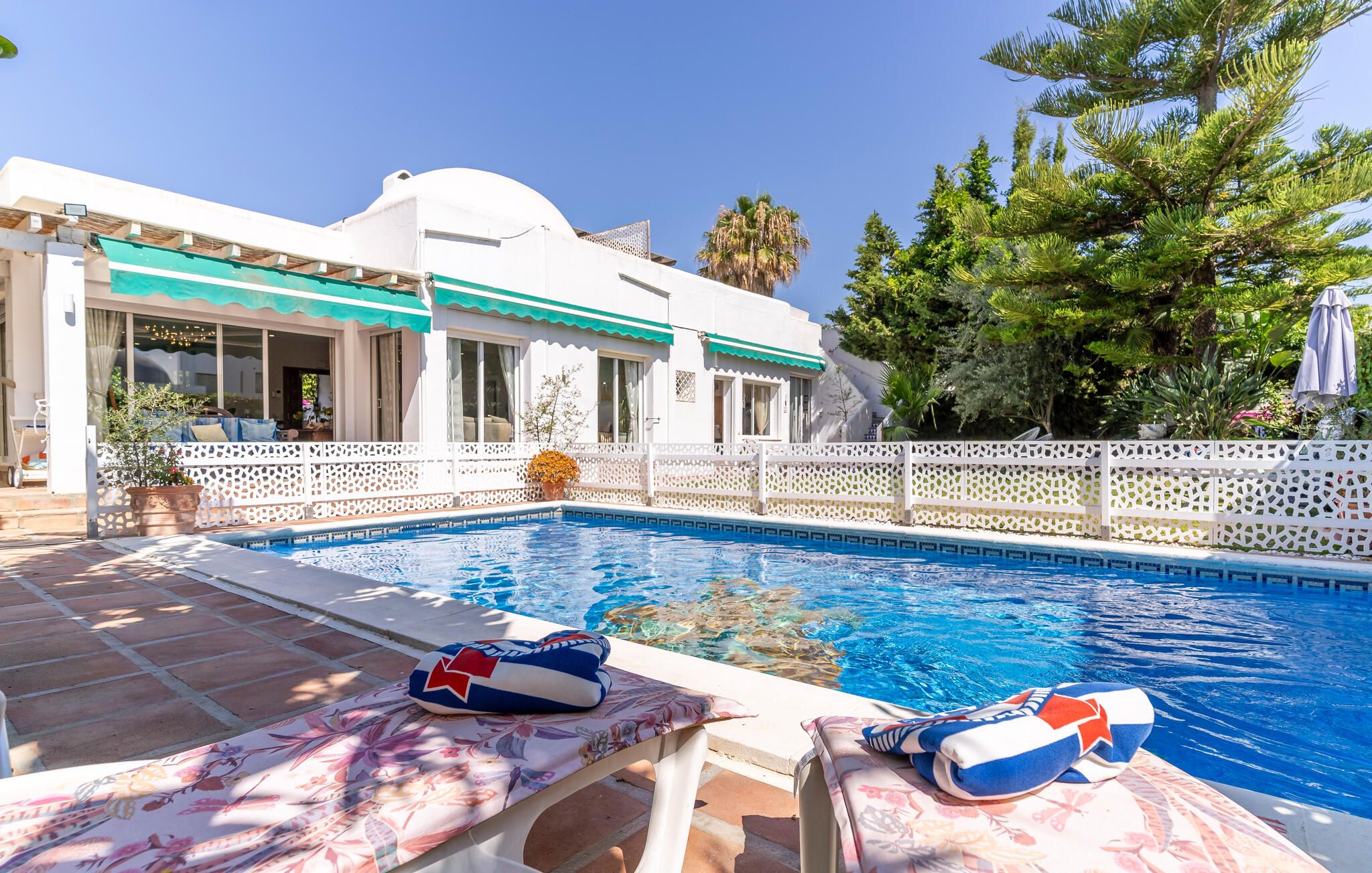 A swimming pool with a white fence, a lounge chair, and a sun umbrella.