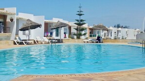 A swimming pool with lounge chairs and thatched umbrellas, surrounded by white buildings with a modern architectural style.