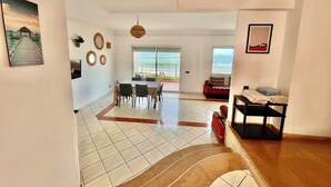 A spacious living area with a dining table and chairs, a sofa, and a view of the ocean through large windows.