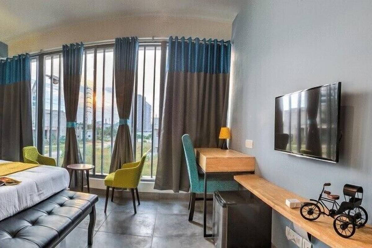 Habitació d'hotel amb llit, escriptori, cadira, banc, televisió i finestra amb cortines.