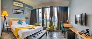 Habitación de hotel con cama, escritorio, silla, banco, televisor y ventana con cortinas.