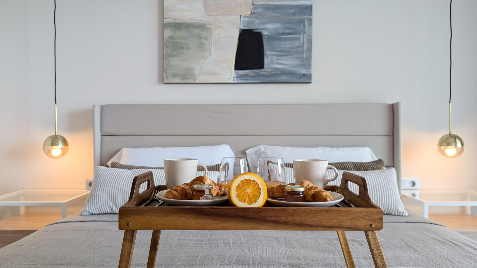 A neatly made bed with a tray of breakfast items, including croissants, orange slices, and coffee cups, in a modern hotel room.