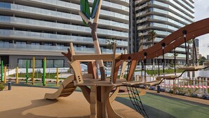 Un parc infantil amb una estructura d'escalada i un edifici al fons amb nombroses balconades.