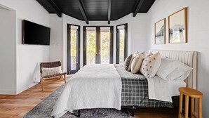 A bedroom with a bed, a chair, a stool, a TV, and a window with a view of trees.