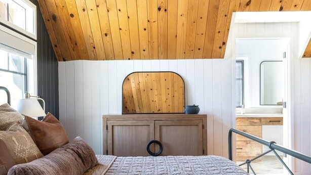 A bedroom with a wooden ceiling, a bed with a grey quilt, a wooden dresser, and a mirror above it.