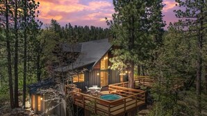 A cabin with a deck, a hot tub, and a small outdoor seating area surrounded by trees.