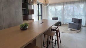 A modern living room with a wooden dining table, two hanging pendant lights, a grey sofa, and a large window with sheer curtains.