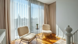 Un salon moderne avec deux fauteuils confortables, une petite table ornée d’une plante et une vue sur le paysage urbain à travers des rideaux légers.