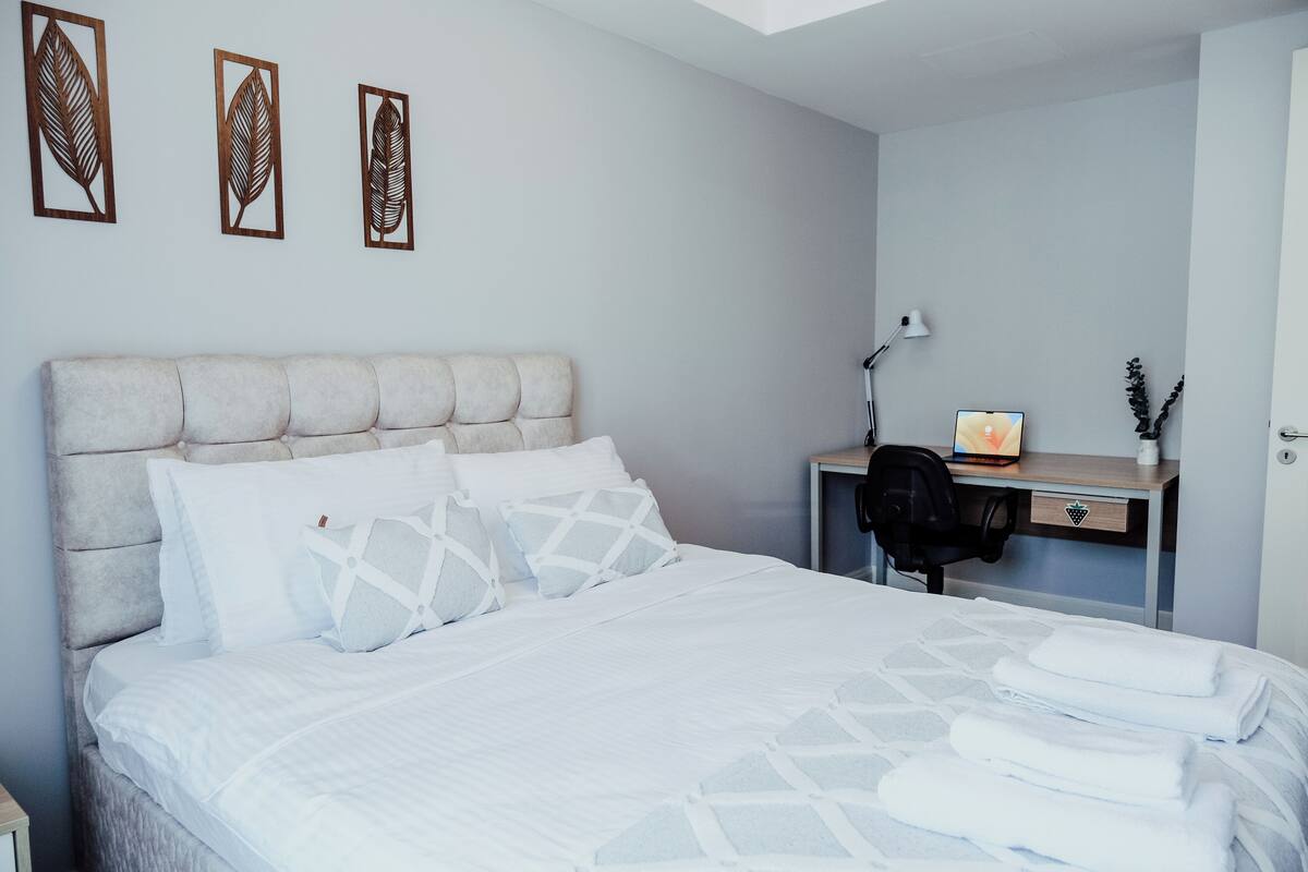 A neatly made bed with white linens and decorative pillows, a desk with a lamp, and a chair.