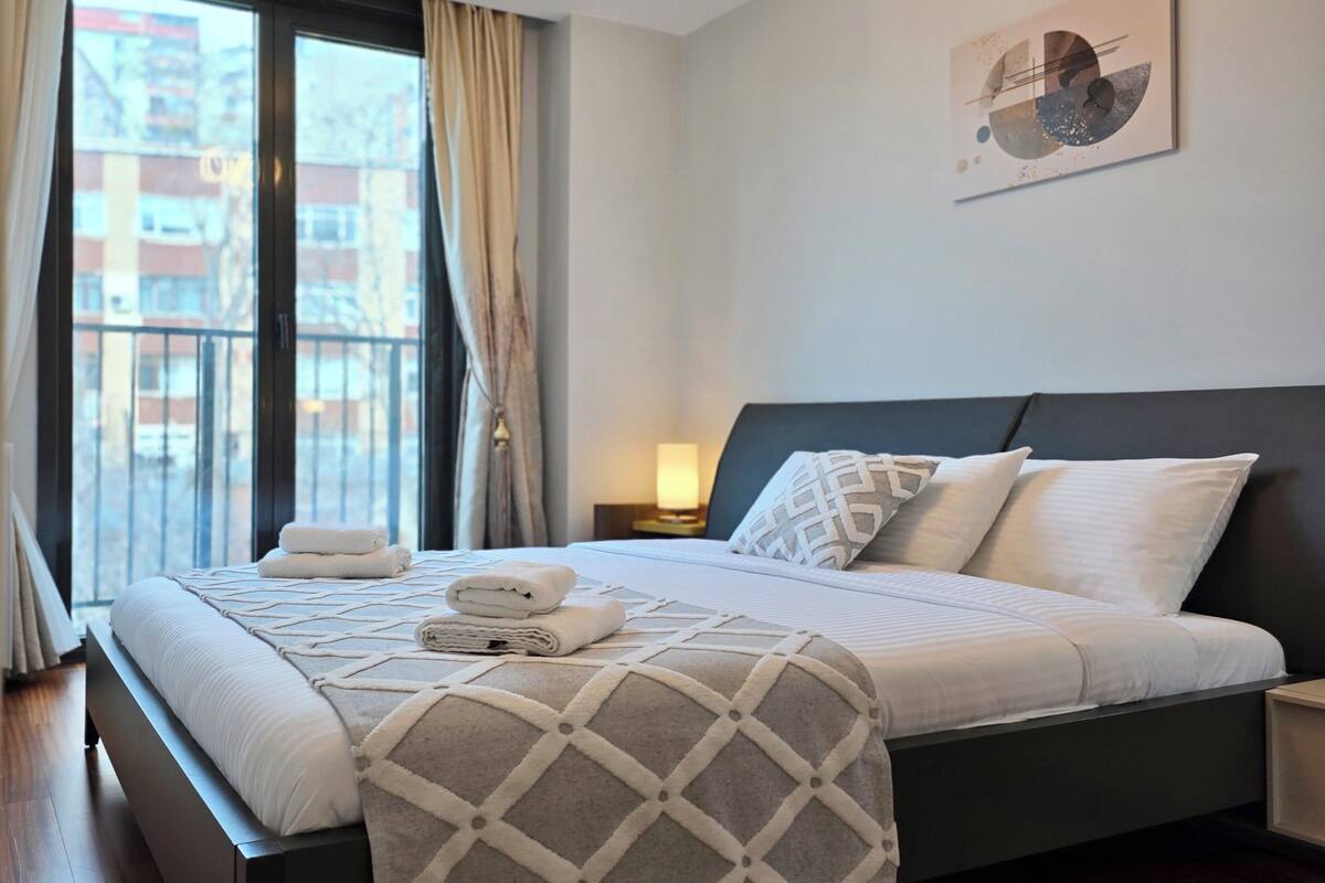 A modern hotel room with a large bed, a window with curtains, a wall-mounted artwork, and a bedside table with a lamp.