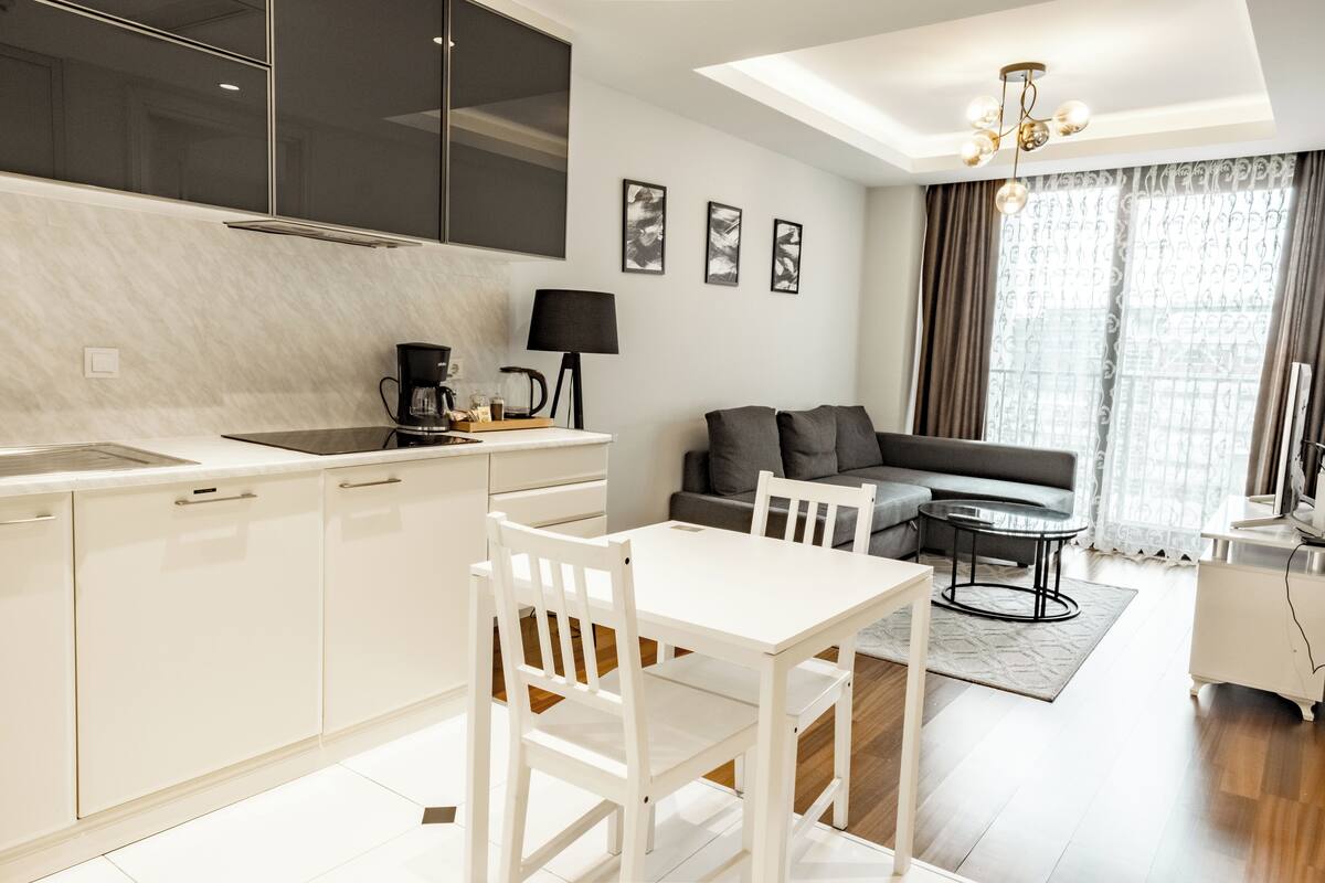 A modern kitchen with white cabinetry, a dining table with chairs, a sofa, and a coffee maker.