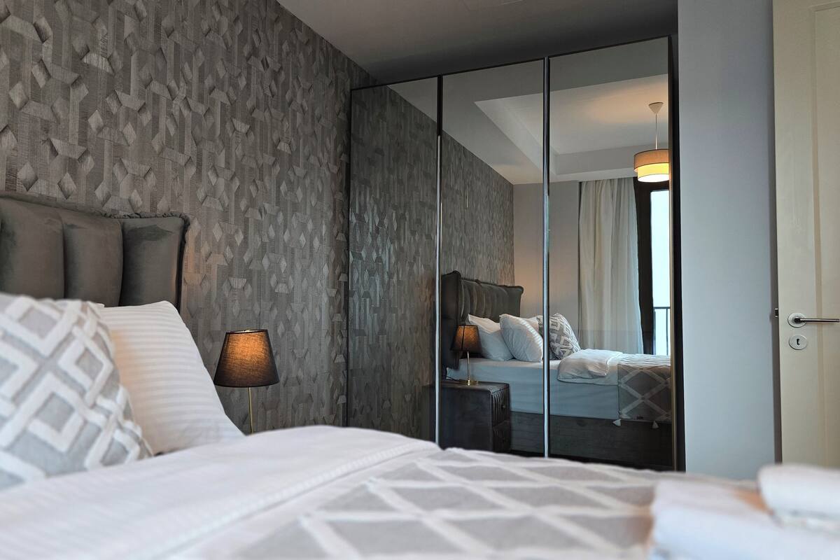 A modern bedroom with a large bed, a nightstand, a lamp, and a wardrobe with a mirror.