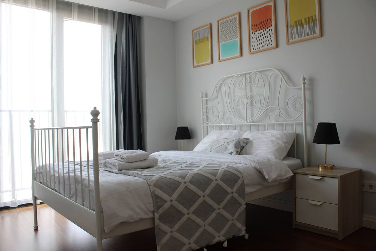 A neatly made bed with a white headboard, a bedside table with a lamp, and a window with sheer curtains.