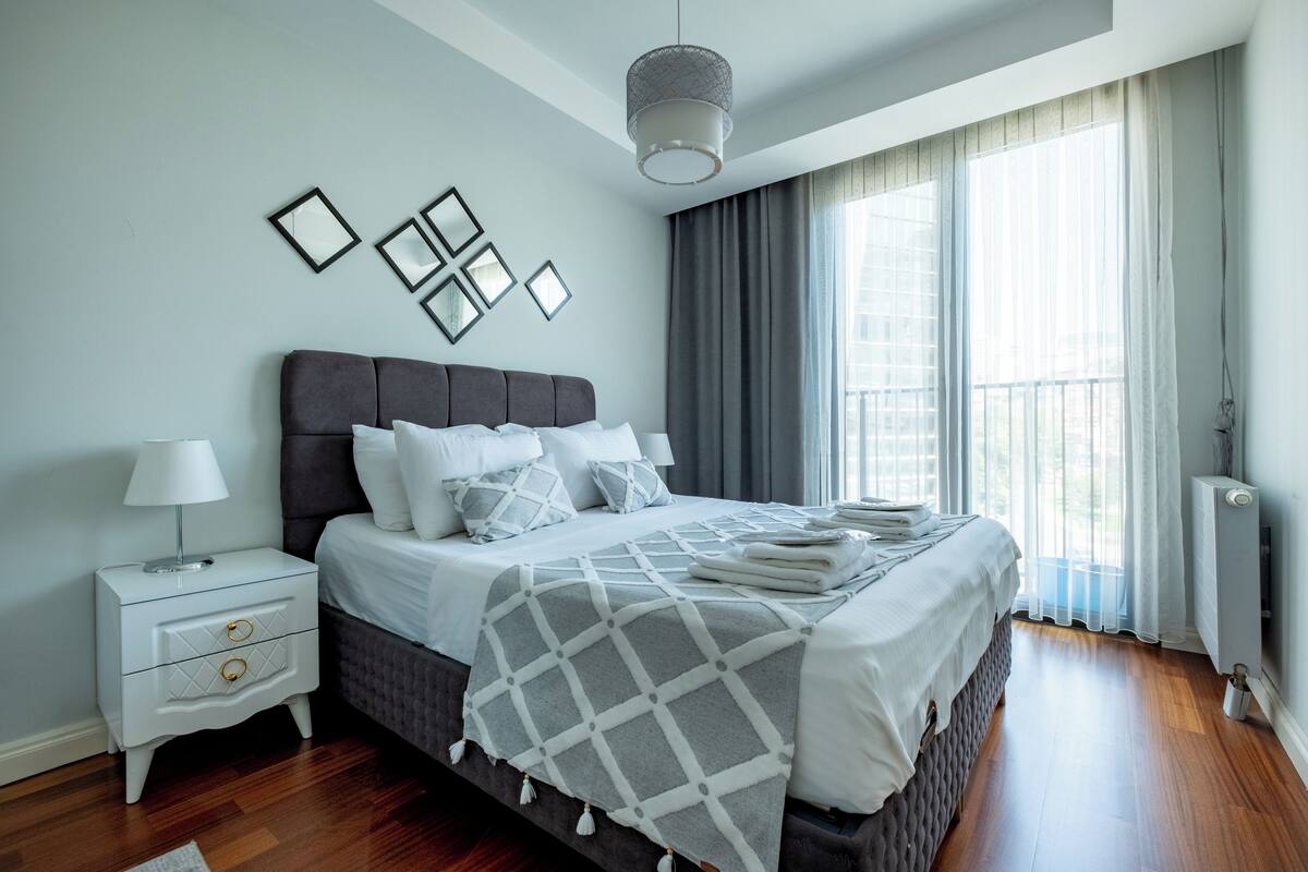 A modern bedroom with a large bed, a nightstand, a lamp, and a window with curtains.