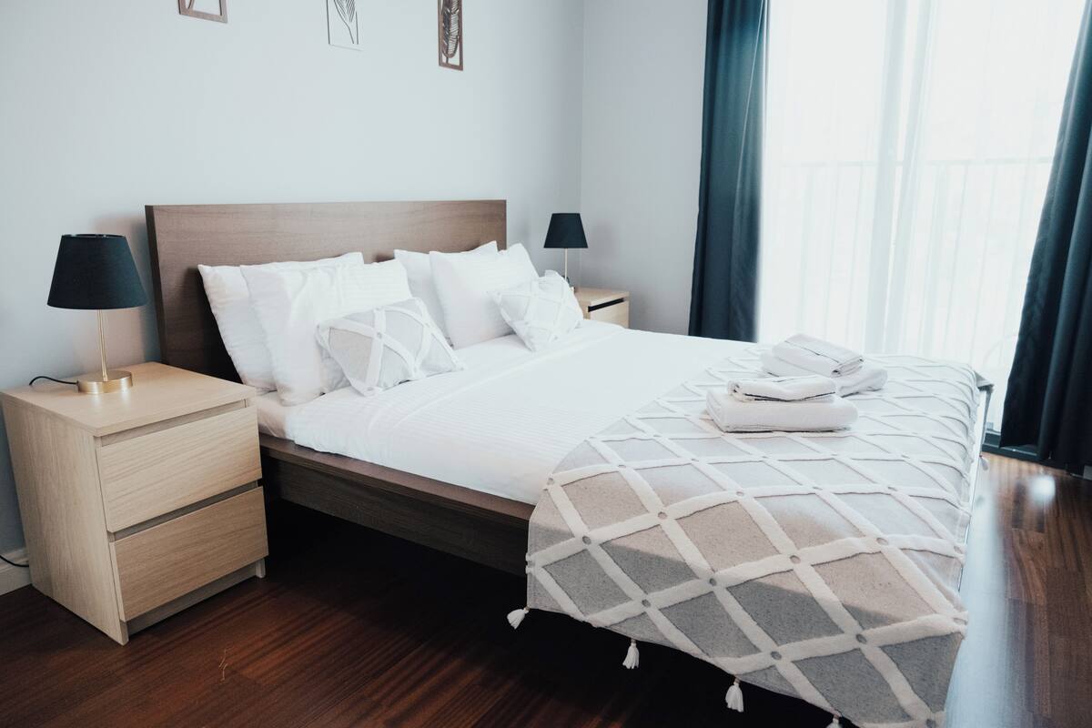 A neatly made bed with a white comforter and pillows, a wooden headboard, and a bedside table with a lamp.