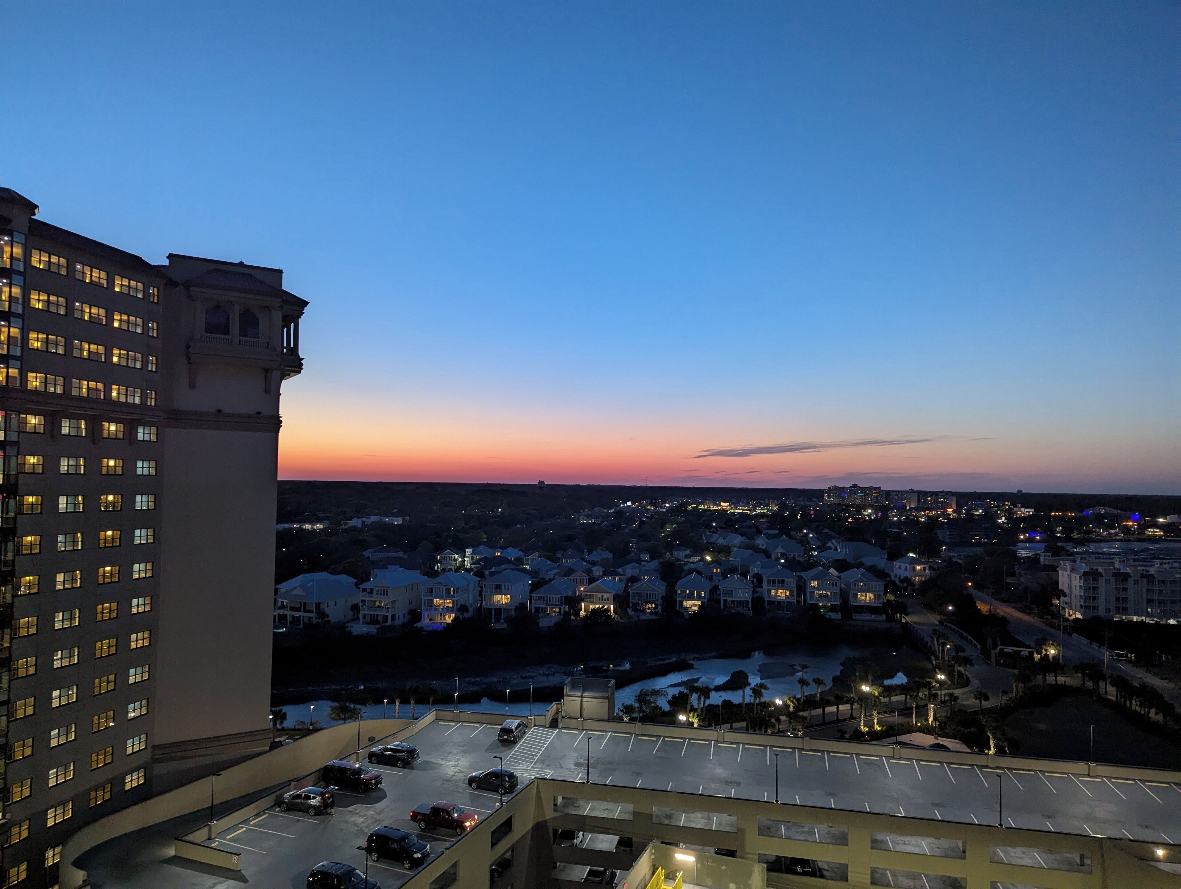 A high-rise hotel with multiple floors, a parking area with several cars, and a view of a residential area with houses and a sunset in the background.