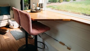 Une cuisine avec un plan de travail en bois, deux tabourets de bar roses, une fenêtre donnant sur un espace verdoyant et une porte noire.