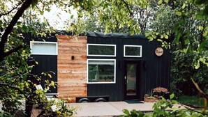 Une maison moderne et compacte, dotée d’une façade noire et de touches de bois, de grandes fenêtres et d’un petit porche à l’avant agrémenté de plantes en pot.