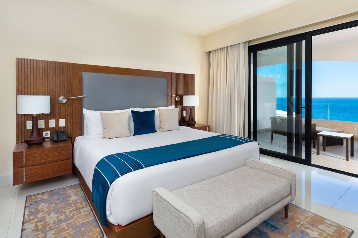Ein Hotelzimmer mit einem großen Bett, einem Nachttisch, einem Schreibtisch, einem Sessel und einem Balkon mit Meerblick.