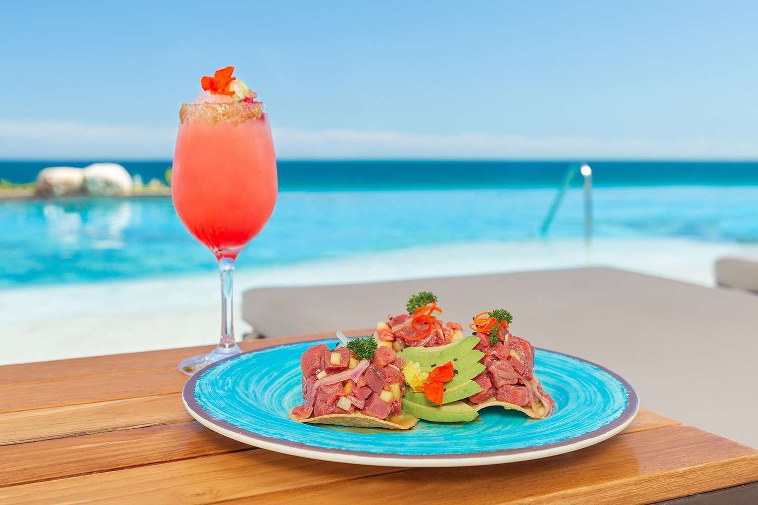 A plate of steak tartare with avocado and vegetables, a pink cocktail, and a view of the ocean.