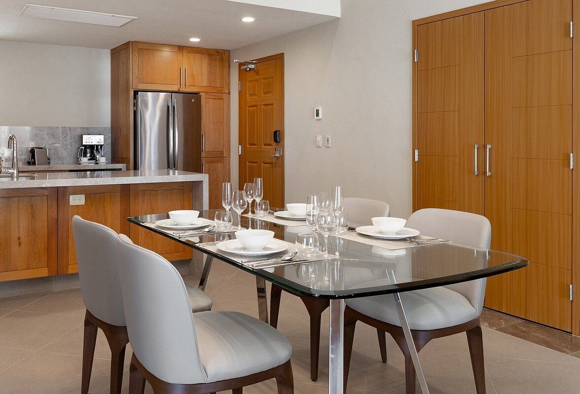 A modern kitchen with wooden cabinets, a stainless steel refrigerator, and a dining area with a glass table set for four.