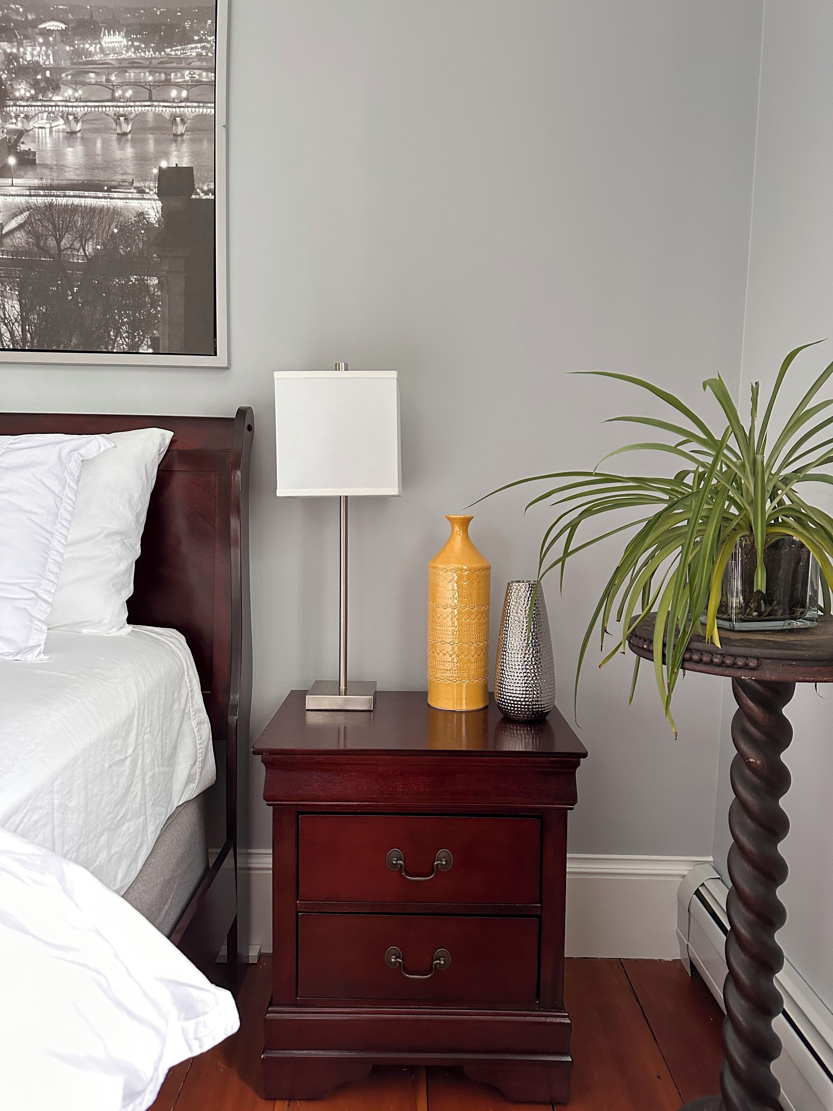 Un dormitorio ordenado con cama, mesita de noche, lámpara, jarrones decorativos y una planta en maceta.